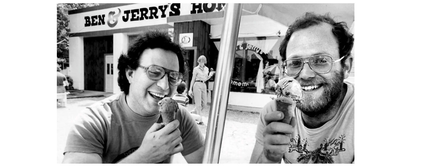 Ben and Jerry sit and eat ice cream cones outside of their founding store. 