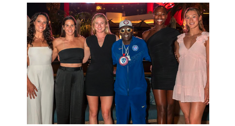 Flavor Flav stands with five members of the US Women’s Water Polo team; he’s dressed in a USA-blue track suit and ball cap, the women are wearing dressier clothing
