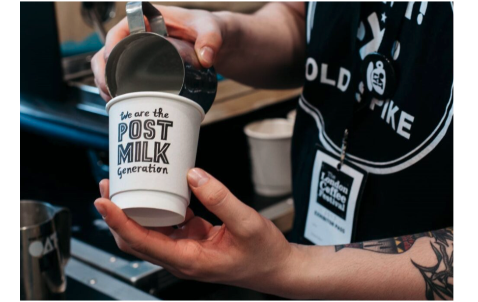 Oatly’s paper cup with the wording, “We are the post milk generation,” is being poured into by a barista with tattoos and a lanyard that readers, “The London Coffee Festival,” around their neck. 