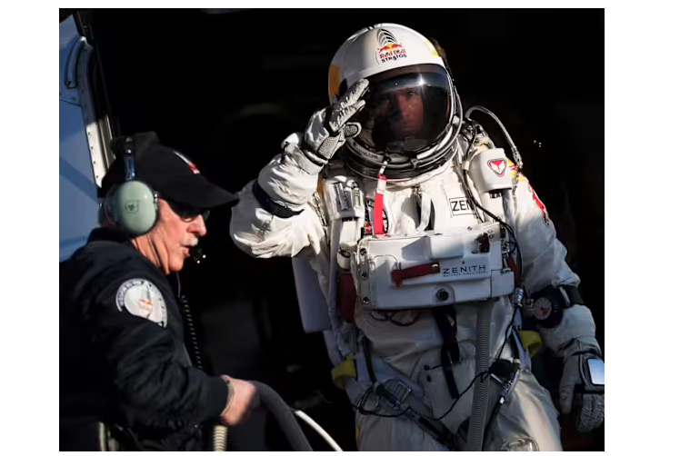 an image of a man in a space suit, saluting with a gloved hand, and in the foreground a white man with a mustache and glasses, with earphones and a microphone and black ball cap. The background is black, making you think of outer space. 