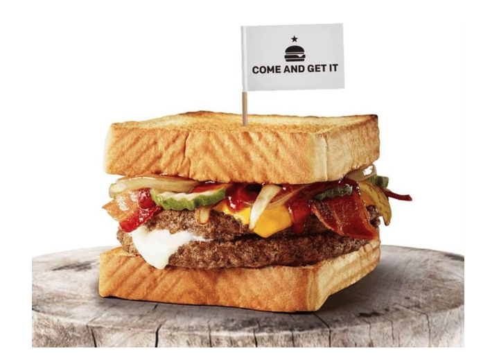 photo of a sandwich with two hamburger patties, cheese, ketchup, bacon, pickles, and onions, on Texas toast, with a toothpick and a tiny flag that reads “come and get it” in the top