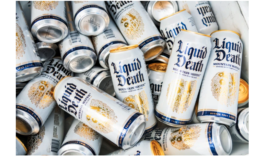 A pile of white aluminum cans, with gold tops and a golden logo and black lettering reading “Liquid Death”