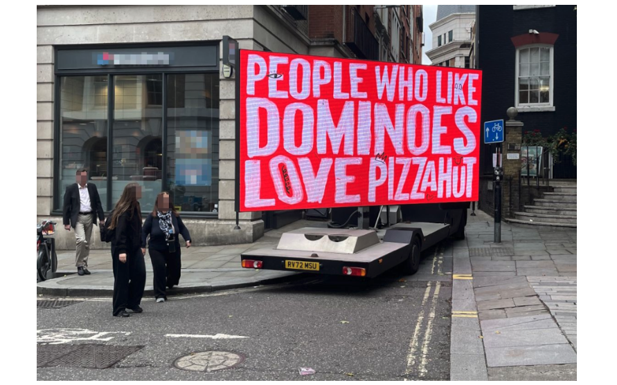 A red billboard on a street corner reads “People who like dominoes love Pizza Hut.” Two women dressed in black with their faces blurred look at the sign. 